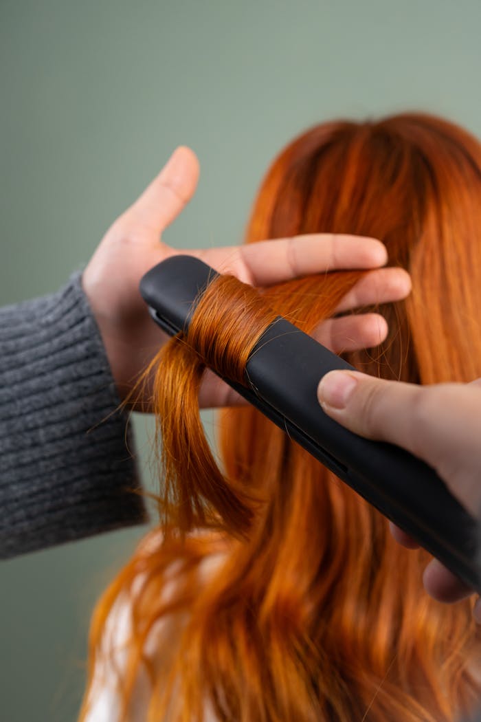 Close-up of red hair being styled with a flat iron indoors.